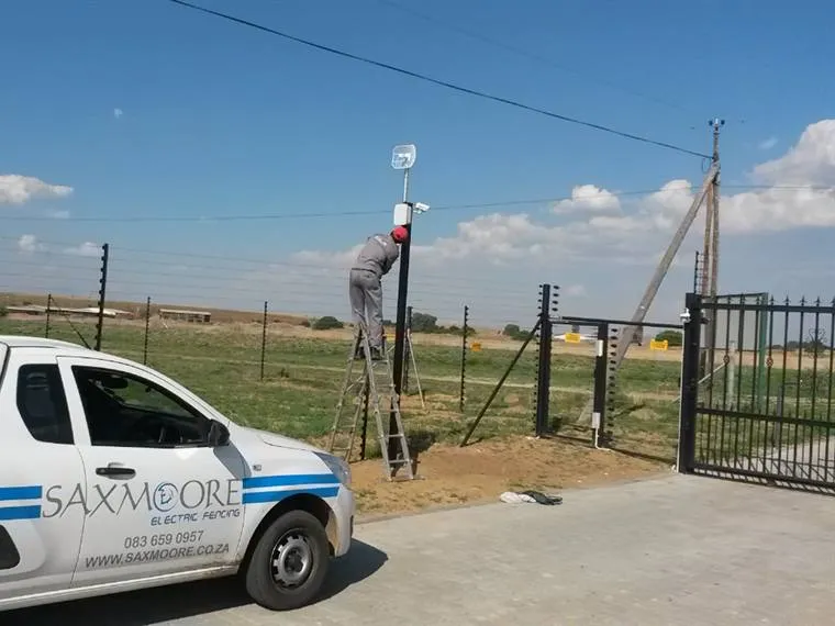 Saxmoore installer on a ladder at a pole beside a freestanding electric fence and gate, company car in front
