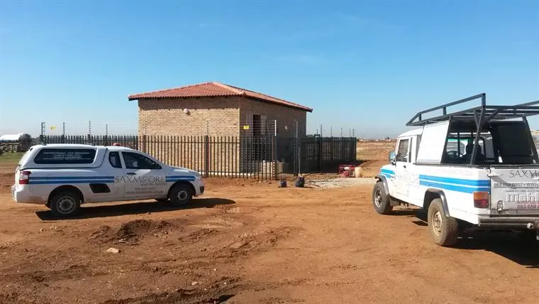 Two Saxmoore vehicles at a palisade and electric fence around a small brick building on open veld